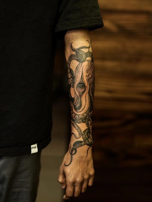 ## Dark Surrealistic Octopus And Winding Tentacle Wraps

Heavy black shading gives this central octopus figure a dense, sculptural weight right on the middle forearm. Winding tentacles creep down toward the wrist and spiral up near the elbow crease. The fleshy texture and detailed suction cups create a high-contrast focal point against the bare skin.

**Fluid, organic shapes wrap the arm naturally exactly like a curved staircase anchors an open floor plan.**

Serpentine designs excel on the forearm because they utilize the full cylindrical canvas without breaking the visual flow. Treat this wrapping layout like draping a heavy velvet runner across a long dining table. You want the thickest part of the subject placed squarely in the middle, allowing the tapered ends to drift cleanly toward the joints.